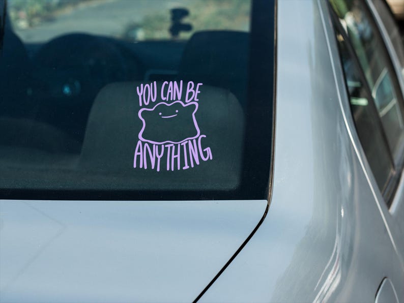 May include: A light purple vinyl decal applied to a car window. The design features a simple, blob-like character with a small face, positioned above and below the text "YOU CAN BE ANYTHING" written in a casual, hand-lettered style font.