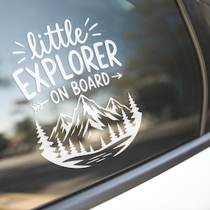 Puede incluir: Adhesivo de vinilo blanco para coche con el texto "little EXPLORER ON BOARD" sobre un diseño de montaña y árboles. El adhesivo está aplicado a una ventana de coche, con una escena exterior borrosa en el fondo.