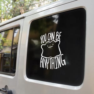 May include: A white vinyl decal applied to a vehicle window, featuring a line-art illustration of a blob-like character with a simple face, accompanied by the text "YOU CAN BE ANYTHING" in a hand-lettered style font above and below the figure.