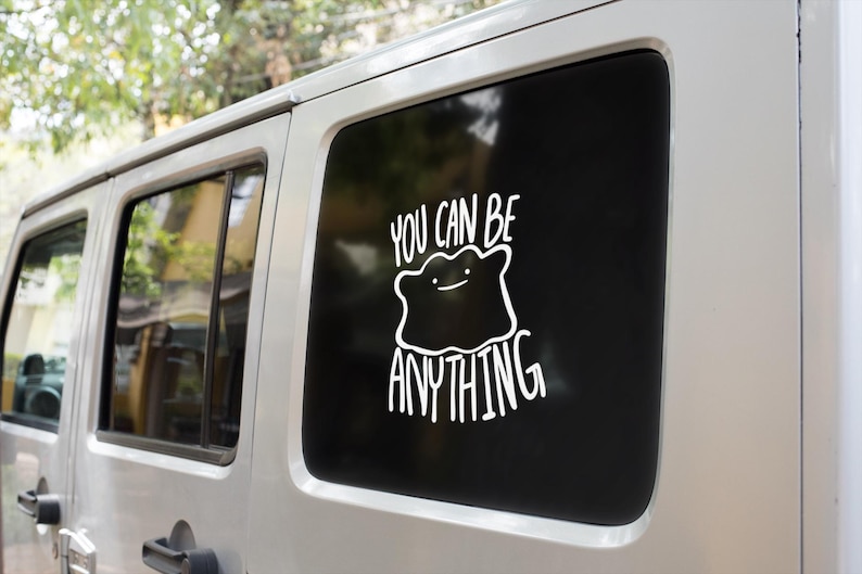 May include: A white vinyl decal applied to a vehicle window, featuring a line-art illustration of a blob-like character with a simple face, accompanied by the text "YOU CAN BE ANYTHING" in a hand-lettered style font above and below the figure.