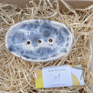 May include: A ceramic soap dish with a blue and white marbled design, featuring three drainage holes. It sits in a cardboard box filled with straw, alongside a bar of Suddy Nora Soap, labeled "Lavender & Rosemary."