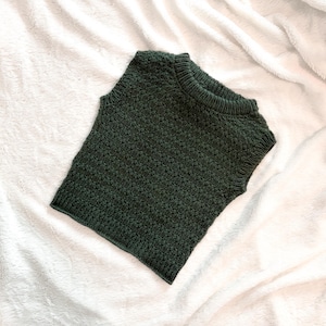 May include: A dark green knitted vest with a crew neck. The sleeveless top is made of a textured knit fabric and is displayed on a white, fluffy surface. The vest is folded neatly.