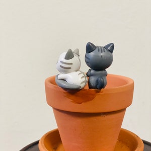 Puede incluir: Dos peque&ntilde;as figuritas de cer&aacute;mica de gatos, una gris y otra blanca, sentadas en el borde de una maceta de terracota. La maceta est&aacute; sobre un platillo marr&oacute;n.