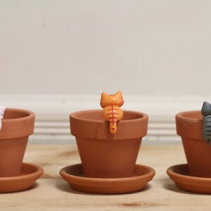 May include: Three terracotta pots with small ceramic cat figurines perched on the rims. The cats are facing away from the viewer, with their tails curled around the pots. The cats are black and white, orange, and gray.