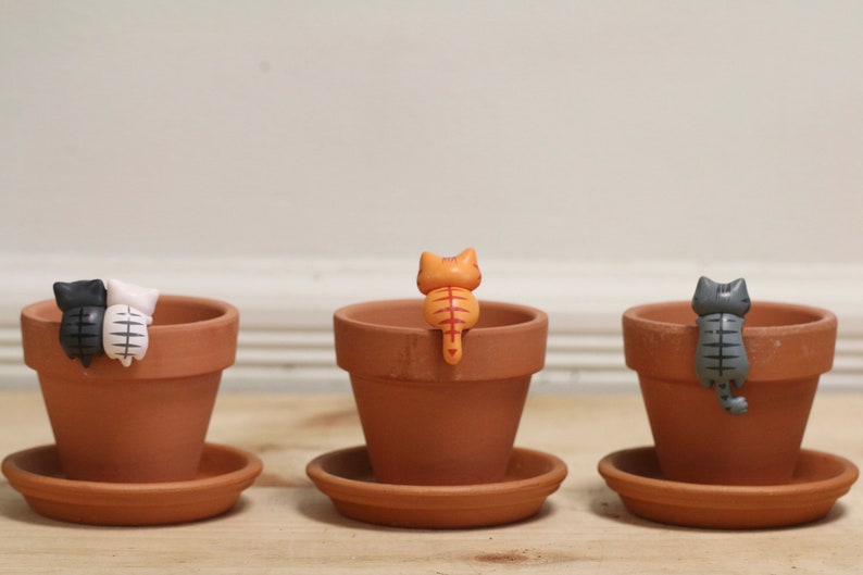 May include: Three terracotta pots with small ceramic cat figurines perched on the rims. The cats are facing away from the viewer, with their tails curled around the pots. The cats are black and white, orange, and gray.