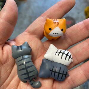 May include: Three small ceramic cat figurines. One orange tabby cat figurine with a white face and a black nose. Two gray tabby cat figurines with black stripes. One gray cat figurine is lying down and the other is standing up.