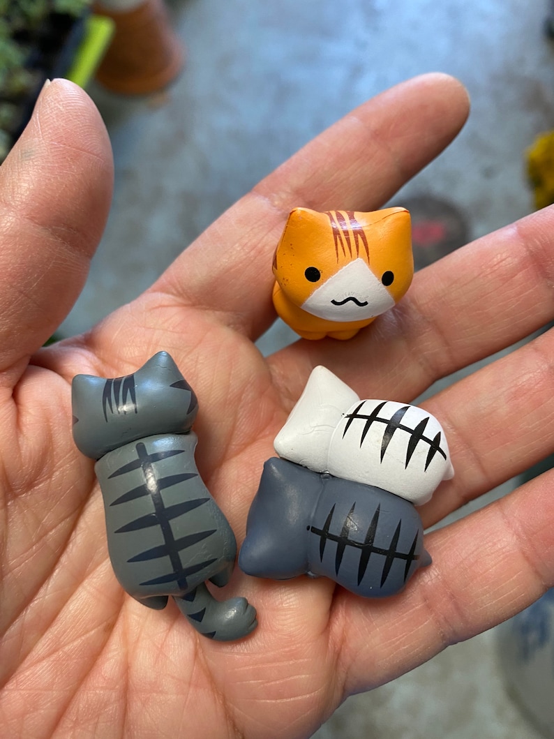 May include: Three small ceramic cat figurines. One orange tabby cat figurine with a white face and a black nose. Two gray tabby cat figurines with black stripes. One gray cat figurine is lying down and the other is standing up.