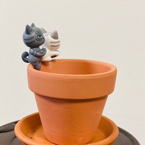 Puede incluir: Una maceta de terracota con un platillo, con dos peque&ntilde;as figuritas de cer&aacute;mica de gatos. Las figuritas de gatos son grises y blancas con rayas negras.