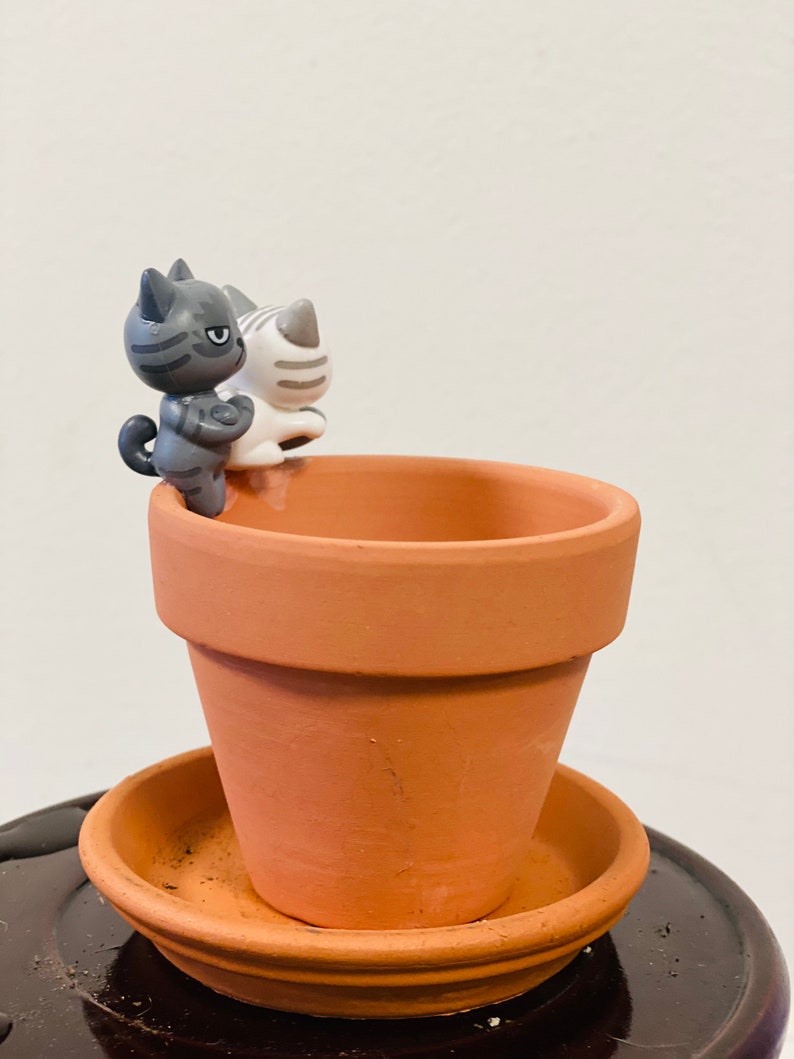 Puede incluir: Una maceta de terracota con un platillo, con dos peque&ntilde;as figuritas de cer&aacute;mica de gatos. Las figuritas de gatos son grises y blancas con rayas negras.