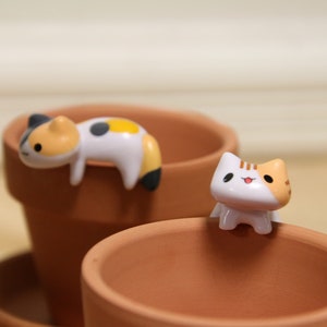 May include: Two small ceramic cat figurines, one white with orange and black markings, the other white with black and yellow markings, sitting on the edge of two terracotta flower pots.