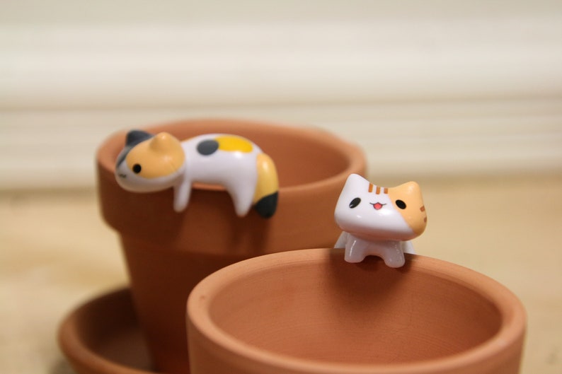 May include: Two small ceramic cat figurines, one white with orange and black markings, the other white with black and yellow markings, sitting on the edge of two terracotta flower pots.