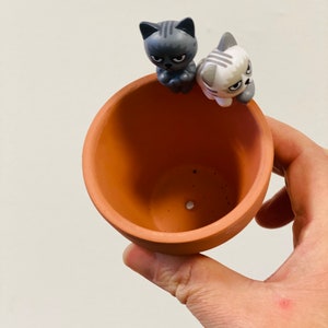 Puede incluir: Una maceta de terracota con dos peque&ntilde;as figuras de gato en el borde. Las figuras de gato son grises y blancas con ojos negros y bigotes.