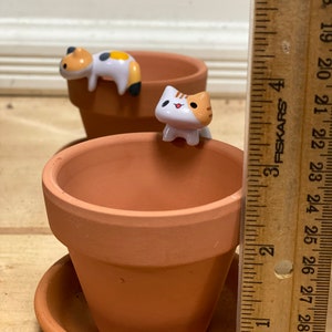 May include: Two terracotta flower pots with cute cartoon cat figurines. The cat figurines are white with orange and black markings. The pots are 3 inches tall.