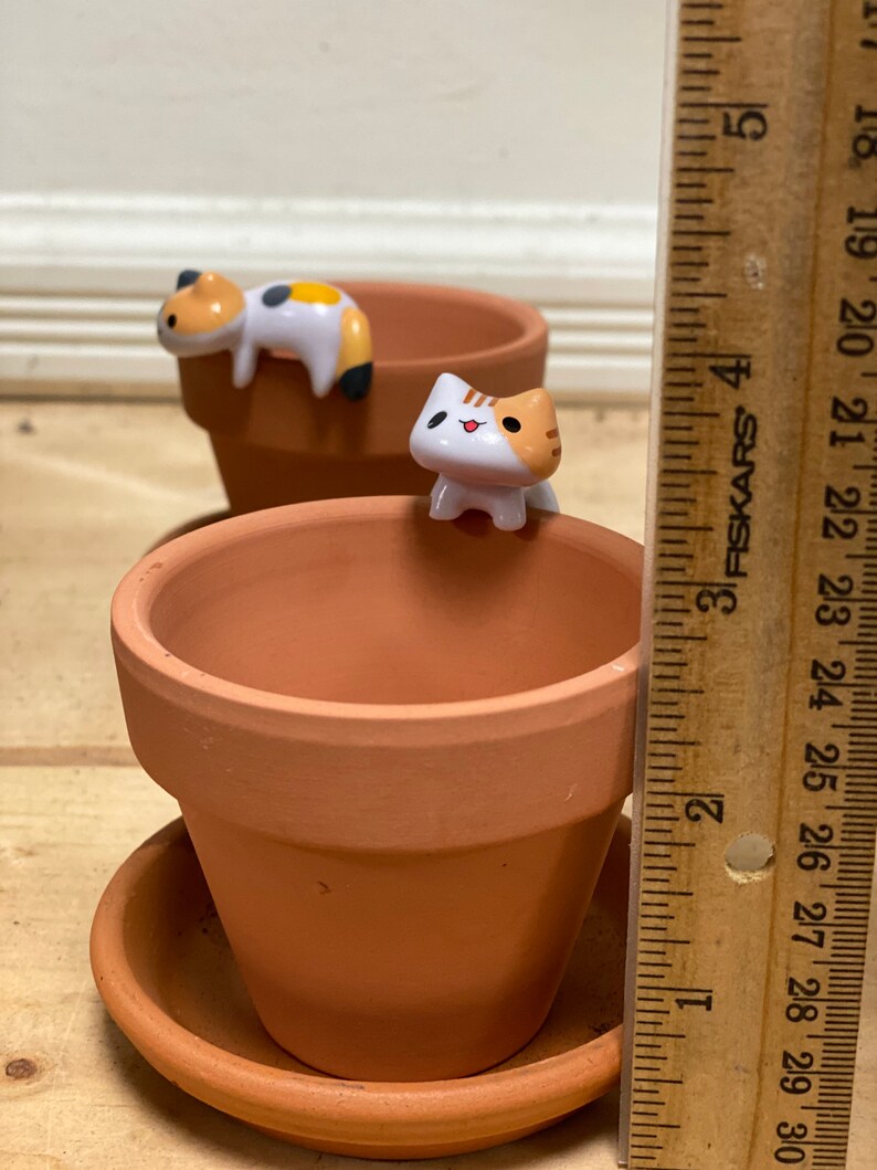 May include: Two terracotta flower pots with cute cartoon cat figurines. The cat figurines are white with orange and black markings. The pots are 3 inches tall.