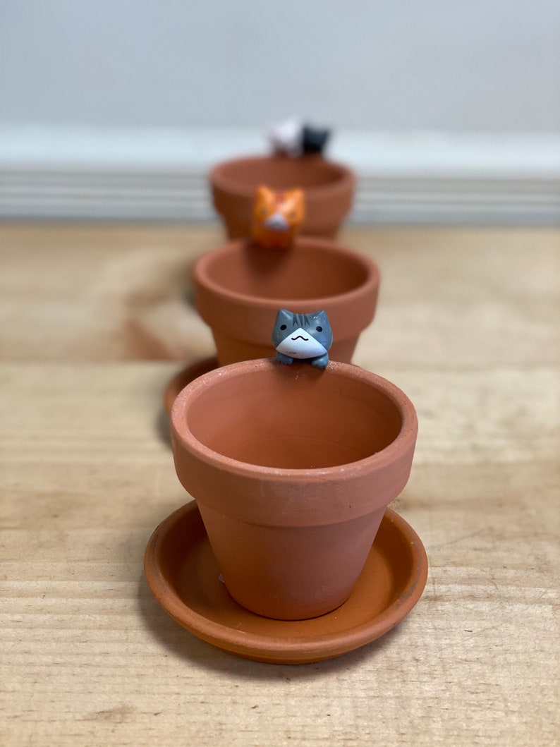 May include: Four terracotta pots with small cat figurines on top. The pots are arranged in a row, with the cat figurines facing the viewer. The pots are all the same size and shape, and they have a brown glaze.