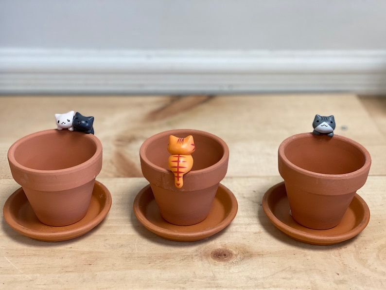 May include: Three terracotta pots with saucers, each featuring a small ceramic cat figurine. The cats are in different colors: white and black, orange and white, and gray and white.