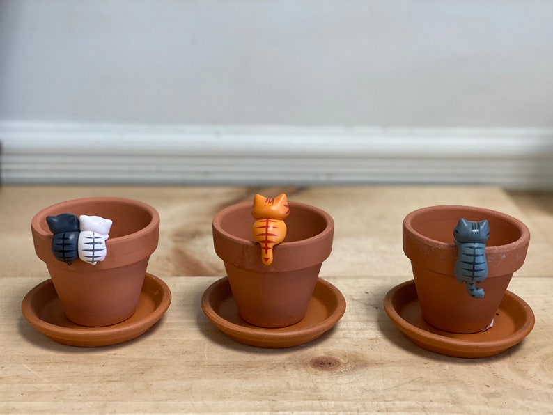 May include: Three terracotta plant pots with decorative cat figurines. The cats are in various colors, including black, white, orange, and gray. The pots are on a wooden surface.