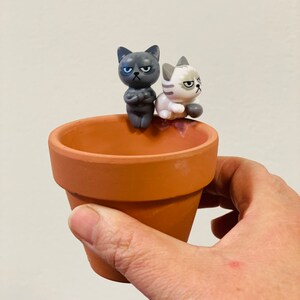Puede incluir: Una maceta de terracota con dos peque&ntilde;as figuras de gatos en la parte superior. Las figuras de gatos son grises y blancas y tienen expresiones de enfado.