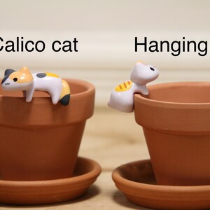 May include: Two terracotta flower pots with decorative cat figurines. One pot has a calico cat figurine laying on the rim, the other has a white cat figurine hanging from the rim. The text "Calico cat" is above the first pot and "Hanging cat" is above the second pot.