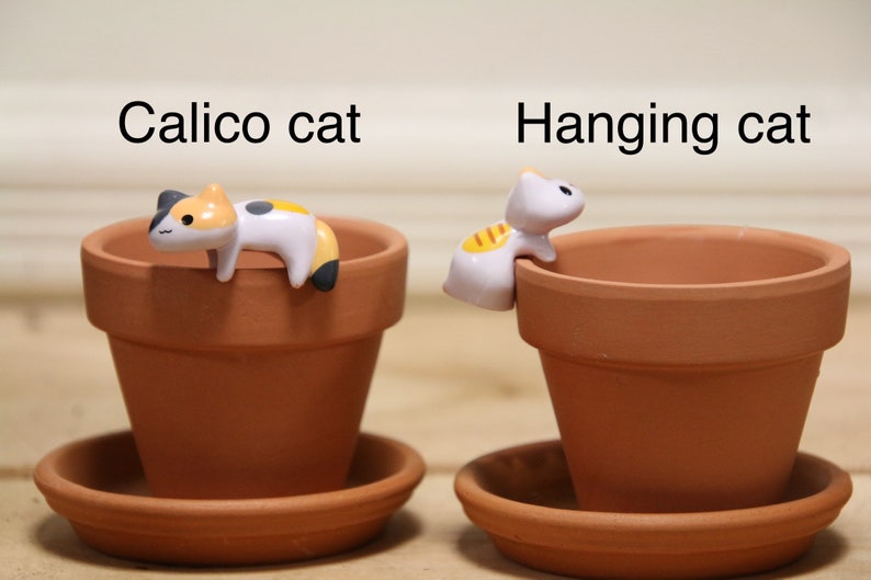 May include: Two terracotta flower pots with decorative cat figurines. One pot has a calico cat figurine laying on the rim, the other has a white cat figurine hanging from the rim. The text "Calico cat" is above the first pot and "Hanging cat" is above the second pot.