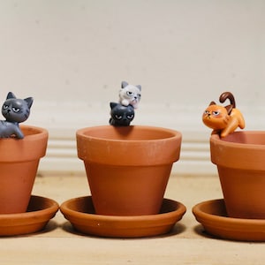 May include: Three terracotta pots with saucers, each featuring a small cat figurine. The cats are in various poses, including sitting, standing, and looking grumpy.