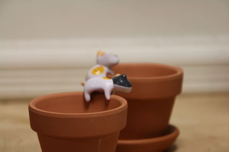 May include: Two small white ceramic cat figurines with yellow and black accents are perched on the rim of a terracotta flower pot. The pot is sitting on a brown surface.