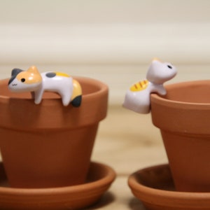 May include: Two terracotta flower pots with cute cat figurines on the rim. The cat figurines are white with black and orange accents. The pots are on matching saucers.