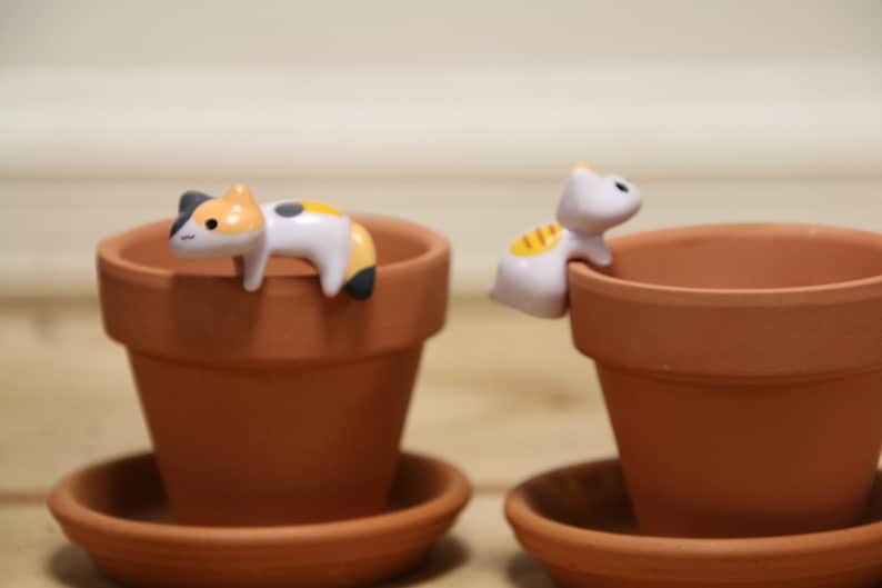 May include: Two terracotta flower pots with cute cat figurines on the rim. The cat figurines are white with black and orange accents. The pots are on matching saucers.