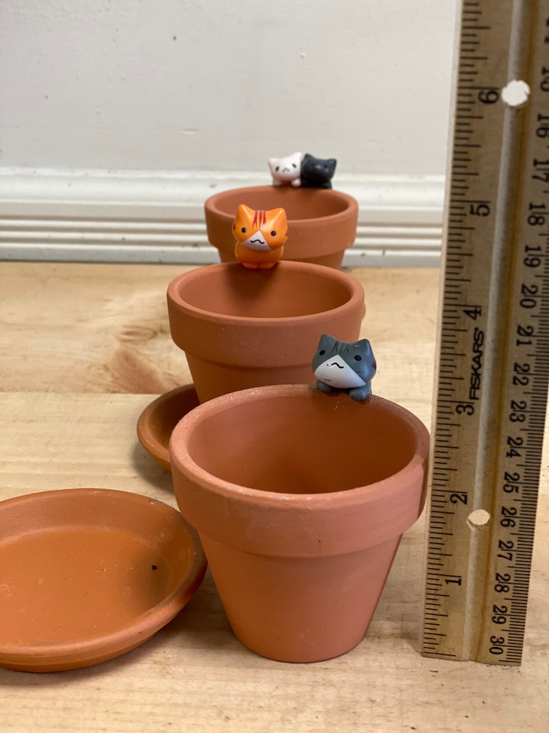 May include: Four terracotta pots with small cat figurines on top. The pots are arranged in a row, with the smallest pot at the front and the largest pot at the back. The pots are all the same color, and the figurines are all different colors.