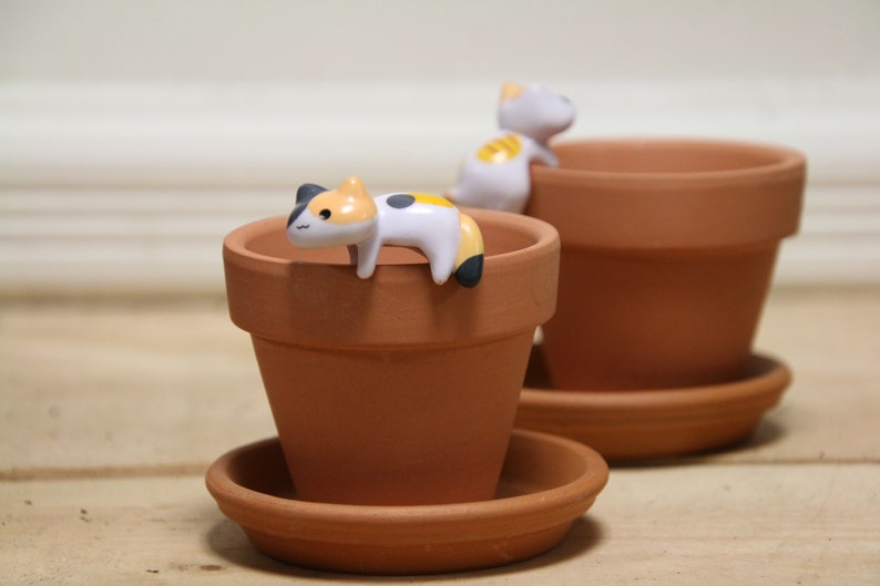 May include: Two terracotta flower pots with small ceramic cat figurines perched on the rims. The cats are white with black and orange markings.