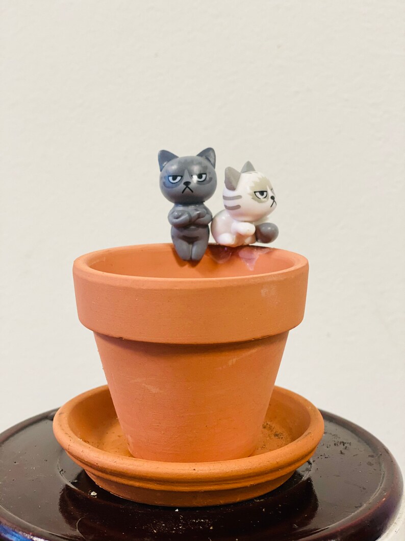 Puede incluir: Dos peque&ntilde;as figuritas de gatos grises y blancos con expresiones gru&ntilde;onas est&aacute;n posadas en el borde de una maceta de terracota con un platillo a juego.
