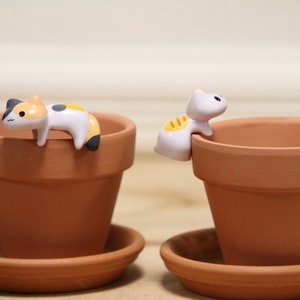 May include: Two terracotta flower pots with ceramic cat figurines. One cat is white with yellow stripes and is perched on the edge of the pot. The other cat is white, black, and orange and is lying on the edge of the pot.