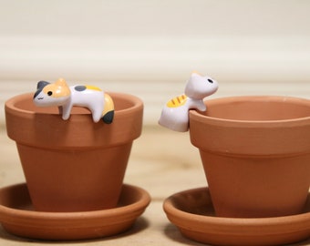 Puede incluir: Dos macetas de terracota con figuritas de gatos de cerámica. Un gato es blanco con rayas amarillas y está encaramado en el borde de la maceta. El otro gato es blanco, negro y naranja y está tumbado en el borde de la maceta.