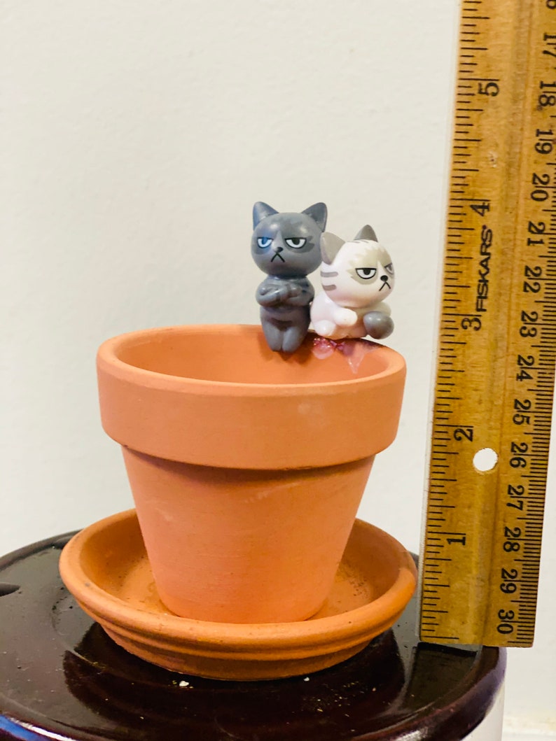 Puede incluir: Dos peque&ntilde;as figuritas de cer&aacute;mica de gatos, una gris y otra blanca, posadas en el borde de una maceta de terracota con un platillo a juego. La maceta mide aproximadamente 7,5 cm de alto.
