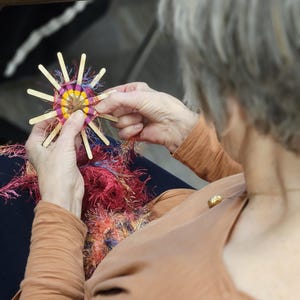 May include: A person weaving yarn on a wooden loom with colorful yarn. The loom is shaped like a star with a circular center.