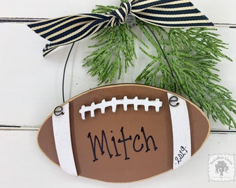 May include: A brown and white football-shaped ornament with the name "Mitch" and the year "2019" written on it. The ornament is decorated with a black and white striped ribbon.