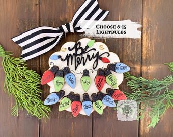 May include: A festive Christmas decoration featuring a white cloud-shaped sign with the words "Be Merry" in black script. Colorful lightbulb ornaments with names are attached. A black and white striped bow and greenery complete the holiday theme.