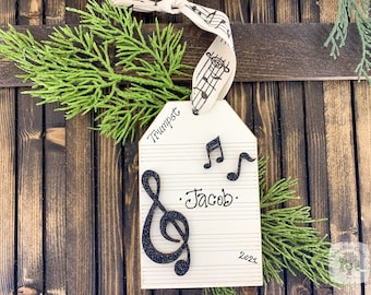 Könnte beinhalten: Ein weißer Holz-Anhänger in Form einer Musiknote mit einem schwarzen Glitzer-Violinschlüssel, zwei schwarzen Noten und dem Text "Trumpet", ".Jacob.", und "2022".