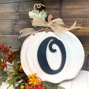 May include: A white wooden pumpkin cutout featuring a large black letter O in the center. The pumpkin is decorated with a burlap ribbon bow at the top, a green leaf accent, and a brown stem. The finish on the pumpkin has a distressed white paint.