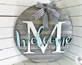 Op de afbeelding: Een ronde houten deurhanger met een grijze houten achtergrond. De letter "M" is uit wit hout gesneden en het woord "welcome" is uit lichtblauw hout gesneden. Een zwart-wit gestreepte strik is aan de bovenkant van de deurhanger bevestigd.