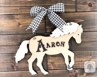May include: A wooden horse cutout with a black and white checkered bow, the name "Aaron" is written on the horse, and the horse is hanging on a wooden wall.