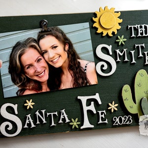 May include: A green wooden picture frame with a photo of two women, a white glitter 'S' and 'F' with the words 'Santa Fe' and 'The Smiths' in white glitter letters, a yellow sun, green cacti, and the year '2023' in white.