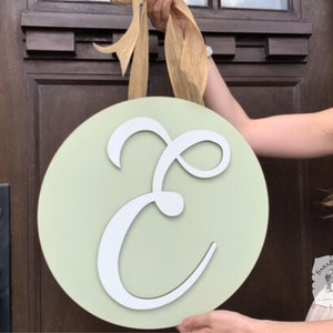 May include: A round, light green wooden door sign with a large white letter "E" in a cursive font. The sign is held up by a tan ribbon bow. The sign is likely for a front door or wall decor.