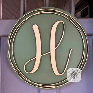 May include: A round wooden sign with a sage green background and a cream-colored letter "H". The letter is in a cursive font. The sign is framed by a thin, double-lined, cream-colored border. The sign is hanging on a purple door.