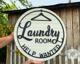 May include: A white round wooden sign with black lettering that reads "Laundry Room Help Wanted". A black hanger is above the text. The sign is held by a hand.