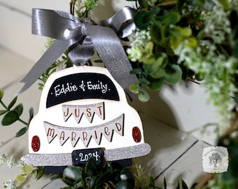 May include: White wooden car ornament with a silver glitter finish. The car has a black chalkboard sign that says "Eddie & Emily. Just Married. 2024."