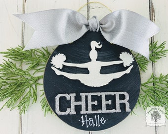 May include: A black and white wooden ornament with a silver glitter finish. The ornament features a silhouette of a cheerleader with pom-poms and the word "CHEER" in silver glitter. The name "Halle" is written below the word "CHEER".