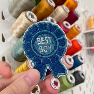 May include: A blue embroidered patch with the text "BEST BOY" in white. The patch is held in front of a collection of colourful thread spools, including yellow, green, and orange. The background is a white pegboard.