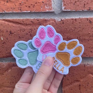 Peut inclure: Trois écussons brodés en forme de pattes de chien, verts, roses et dorés. Chaque écusson a un fond blanc et un nom brodé dans une couleur assortie. Les écussons sont tenus devant un mur de briques.