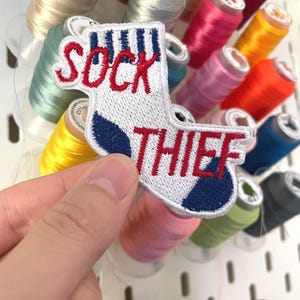 Könnte beinhalten: Gestickter Aufnäher in Form einer weißen Socke mit blauen Akzenten und den roten Worten "SOCK THIEF". Der Aufnäher wird vor bunten Garnrollen gehalten.